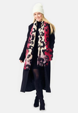 Schwarzer langer Mantel, gestrickte weiße Mütze und mehrfarbiger Leopardenmuster-Schal, der von Rot nach Beige übergeht. Knielange schwarze Stiefel vervollständigen das Outfit.