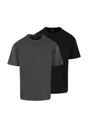 Jeune homme portant un t-shirt noir uni et un short gris clair, debout devant un fond gris clair uni.