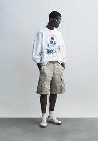 Jonge man met een wit sweatshirt met palmboomprint, beige cargoshort, witte sokken, sneakers en een zonnebril, staand met handen in de zakken.