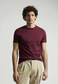 Camiseta de manga corta burdeos en una suave mezcla de algodón, con escote redondo y un sutil detalle del logo en el pecho. Combinada con pantalones beige.