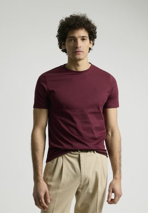 Camiseta de manga corta burdeos en una suave mezcla de algodón, con escote redondo y un sutil detalle del logo en el pecho. Combinada con pantalones beige.