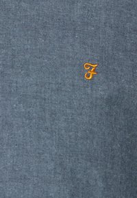 Tissu bleu avec une petite lettre brodée orange "F" près du centre.