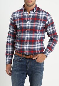 Chemise à carreaux boutonnée en bleu marine, rouge et blanc ; en coton ; manches longues ; avec un col et des boutons sur le devant ; portée avec un jean en denim.