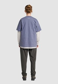 Fioletowa bawełniana koszulka z krótkim rękawem, nałożona na szary T-shirt z długim rękawem. Luźne czarne spodnie i ciemne obuwie, widok z tyłu.