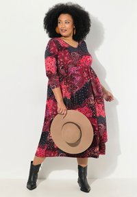 Punainen ja musta kuviollinen mekko, jossa on V-kaula-aukko, kolmannelta neljännekseltä olevat hihat, kerätty vyötärö ja virtaava hame. Malli pitää laajapohjaista hattua.
