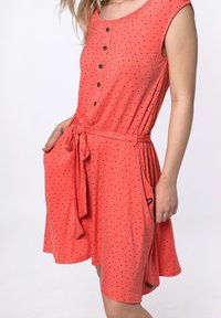 Korálové šaty s kulatým výstřihem, krátkými rukávy, knoflíky po celé délce a páskem na stažení v pase. Obsahují kapsy a malý černý srdcový vzor.