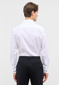 Camicia bianca a maniche lunghe con una texture liscia, polsini abbottonati e colletto standard, abbinata a pantaloni neri con tasche posteriori.