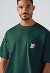 Mand med kort skæg iført en mørkegrøn T-shirt med brystlomme samt et lille rødt og hvidt mærke på lommen, neutral baggrund.