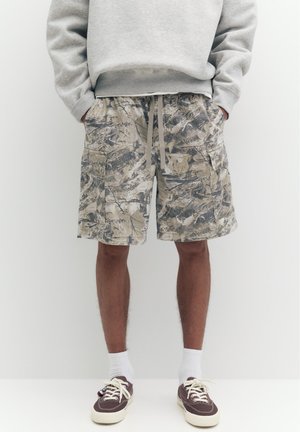 Mann trägt graues Sweatshirt, abstrakte Camouflage-Cargoshorts mit Kordelzug, weiße Socken und dunkelbraune Leinenschuhe, steht vor weißem Hintergrund.