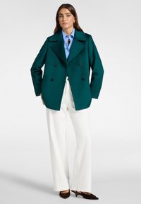 Donna che indossa un cappotto doppiopetto verde scuro, una camicia azzurro chiaro, pantaloni bianchi a gamba larga e scarpe con tacco marroni scuri, in piedi davanti a uno sfondo bianco.