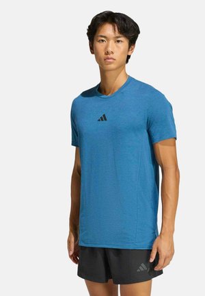 Giovane uomo in piedi che indossa una maglietta Adidas blu e pantaloncini Adidas neri su uno sfondo bianco semplice.