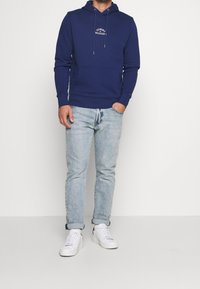 Felpa blu navy con cappuccio, cordini e tasca frontale, abbinata a jeans azzurri e sneakers bianche. Il materiale sembra essere una miscela di cotone.