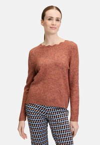 Rustfarbener Pullover aus strukturiertem Strickstoff, mit einem gewellten Ausschnitt und gerippten Bündchen. Kombiniert mit gemusterten Hosen.