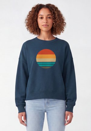 Femme aux cheveux bouclés portant un sweat-shirt bleu marine avec un motif rayé circulaire aux couleurs chaudes et froides, associé à un jean bleu clair.