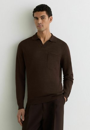 Férfi sötétbarna, hosszú ujjú kötött galléros pólóban, mellkasi zsebbel, világos fal előtt áll, kezét a zsebében tartja, semleges arckifejezéssel.