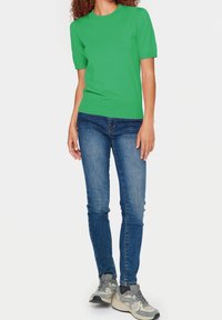 Zelený úpletový svetr s krátkým rukávem a kulatým výstřihem, kombinovaný s modrými slim-fit džíny a šedými teniskami s jemnými akcenty.