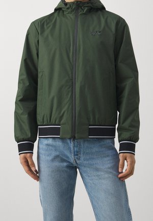 Homme portant une veste à capuche zippée verte avec des poignets et une ceinture rayés noirs et blancs, associée à un jean bleu.