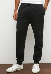 Sorte sweatpants med elastisk talje, smalle manchetter og sidelommer, parret med hvide sneakers på et trægulv.