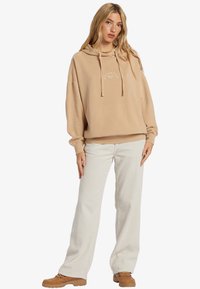 Sudadera con capucha color beige claro, con cordones y logo blanco, combinada con pantalones de pana anchos color blanco y zapatos marrones.