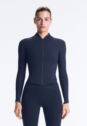 Femeie cu păr închis la culoare, purtând o jachetă sport cu fermoar și mâneci lungi, de culoare bleumarin, și colanți asortați, stând cu fața către înainte pe un fundal alb simplu.