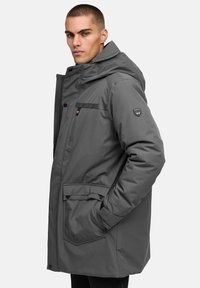 Grå isolerad parkas med huva, ficka med dragkedja på bröstet, stora framfickor och en logo på ärmen. Slät yta, medellång design.