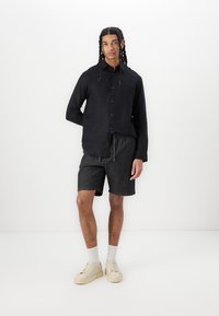 Chemise boutonnée en lin noir, shorts foncés avec cordon de serrage, baskets crème et chaussettes blanches. Le modèle se tient devant un fond blanc uni.