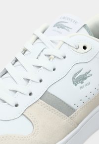 Λευκό δερμάτινο αθλητικό παπούτσι με σουέντ λεπτομέρειες, γκρι πλευρικές ρίγες και διάτρητες λεπτομέρειες. Διαθέτει λογότυπο Lacoste και επίπεδα λευκά κορδόνια.