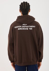 Bruine oversized hoodie met een trekkoord in de capuchon. Witte tekst met "BERLIN DESIGN DEPARTMENT ARCHIVE ‘95" op de achterkant. Geribbelde manchetten en zoom.
