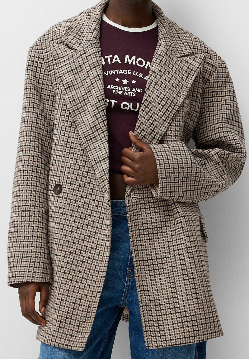 Personne portant un manteau à carreaux beige et marron par-dessus un haut court bordeaux avec un texte blanc et un jean bleu.