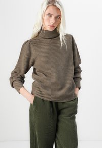 Brun ribbad tröja med hög polokrage och samlade ärmar, matchad med gröna vida byxor i kordfloret. Slät textur, avslappnad passform.