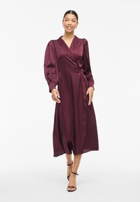 Burgundy satin wrap dress with puffed sleeves and a sash detail. Features a mid-length hem and smooth texture. Black heeled sandals.