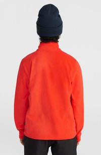 Rode fleece jas met hoge kraag, lange mouwen en een gladde textuur, gecombineerd met een marineblauwe gebreide beanie. Achteraanzicht.