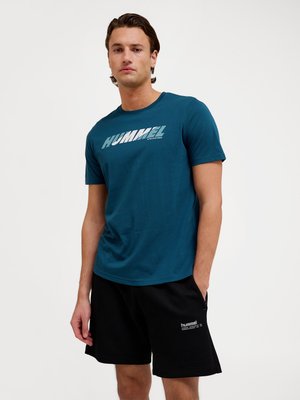 Ung mand stående med hænderne i lommerne iført blå Hummel t-shirt og sorte Hummel shorts mod en ensfarvet hvid baggrund.