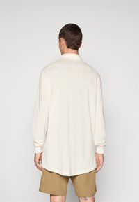 Langermet polo skjorte i off-white stoff med knappet krage, drop shoulder-sømmer og buet hem. Båret med beige shorts.