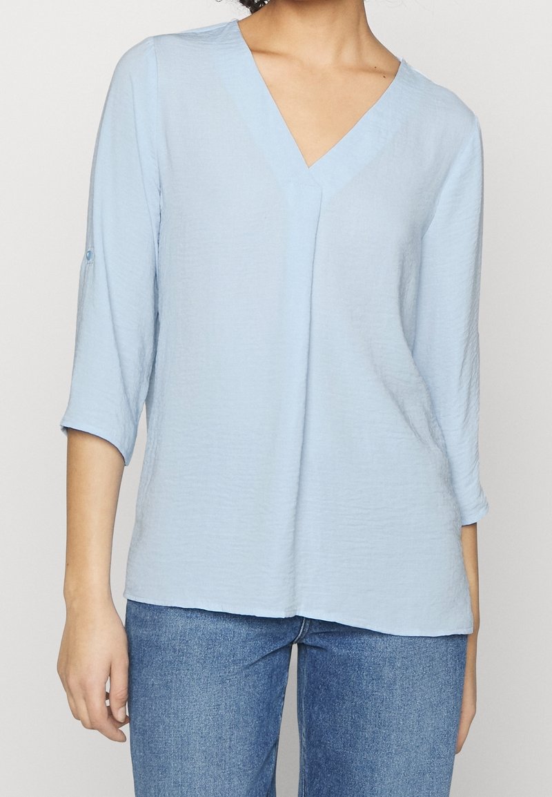Blusa azzurra chiara con scollo a V, maniche a tre quarti, tessuto texturizzato e vestibilità rilassata, abbinata a jeans blu.