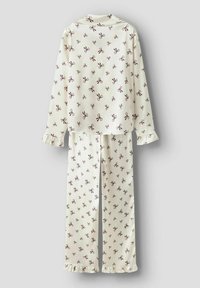 Crème-kleurige pyjama set met lange mouwen en bruine strikpatronen, met ruches aan de mouwen en broekspijpen, weergegeven op een lichtgrijze achtergrond.