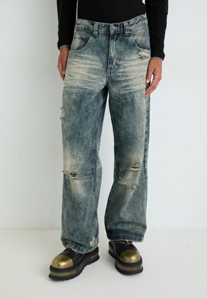 Faded blue denim jeans with a loose fit, distressed detailing, and visible rips. Worn with black and gold platform shoes.