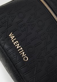 Crna torba od teksturirane kože s uzorkom podignutog loga, sa zlatnim patentnim zatvaračem i zlatno ugraviranim imenom branda "VALENTINO."