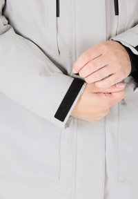 Hellgraue wasserdichte Jacke mit einem schwarzen Klettverschluss am Bund und einem Frontreißverschluss, mit glatter Textur und minimalistischem Design.
