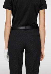 Svart golfoutfit med en kortärmad skjorta och skräddarsydda byxor i en subtil mönstrad textur. Inkluderar ett svart bälte och en vit golfhandske.