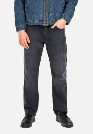 Mann trägt eine blaue Jeansjacke, schwarze Jeans, schwarze Schnürschuhe, mit einer Hand in der Tasche und der anderen locker neben dem Körper.