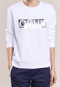Persona che indossa un felpa bianca con un logo rettangolare argento lucido e il testo "HUGO" rovesciato, abbinata a pantaloni blu navy con strisce bianche.