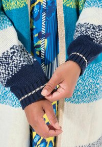Strickjacke mit blauen, grünen und cremefarbenen Mustern, mit gerippten marineblauen Bündchen mit einem cremefarbenen Streifen. Floral bedrucktes Kleid darunter sichtbar.