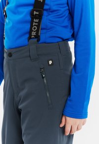 Grijze waterdichte broek met een ritssluiting zak en zwart logo-patch, gedragen over een blauwe long sleeve shirt met blauwe mouwen.