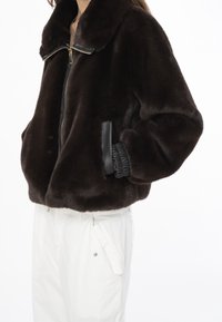 Brązowa sztuczna futrzana kurtka z czarnym zamkiem, kołnierzem i bocznymi kieszeniami. Ozdobiona elastycznymi akcentami przy rękawach, tworzy puszystą sylwetkę.