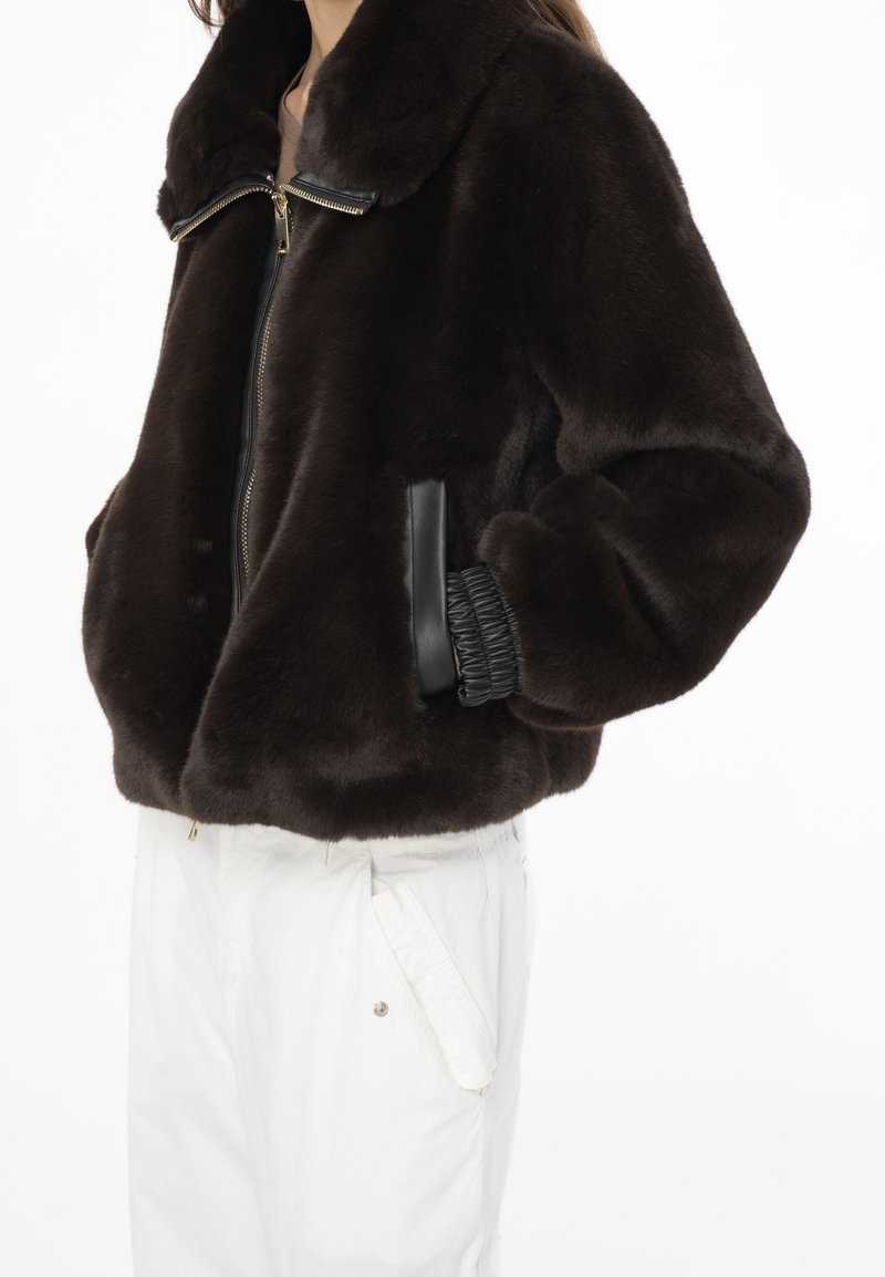 Brązowa sztuczna futrzana kurtka z czarnym zamkiem, kołnierzem i bocznymi kieszeniami. Ozdobiona elastycznymi akcentami przy rękawach, tworzy puszystą sylwetkę.