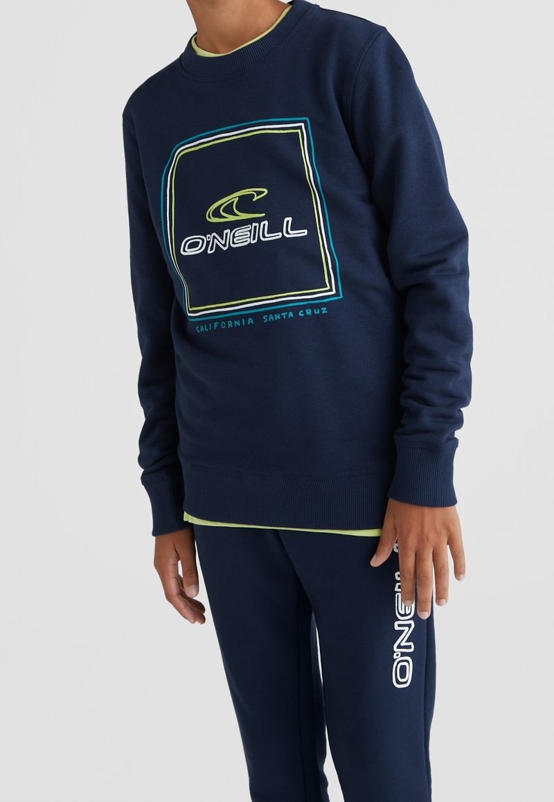 Personne portant un sweat-shirt bleu marine O'Neill et un pantalon assorti avec les logos de la marque, debout devant un fond clair uni.