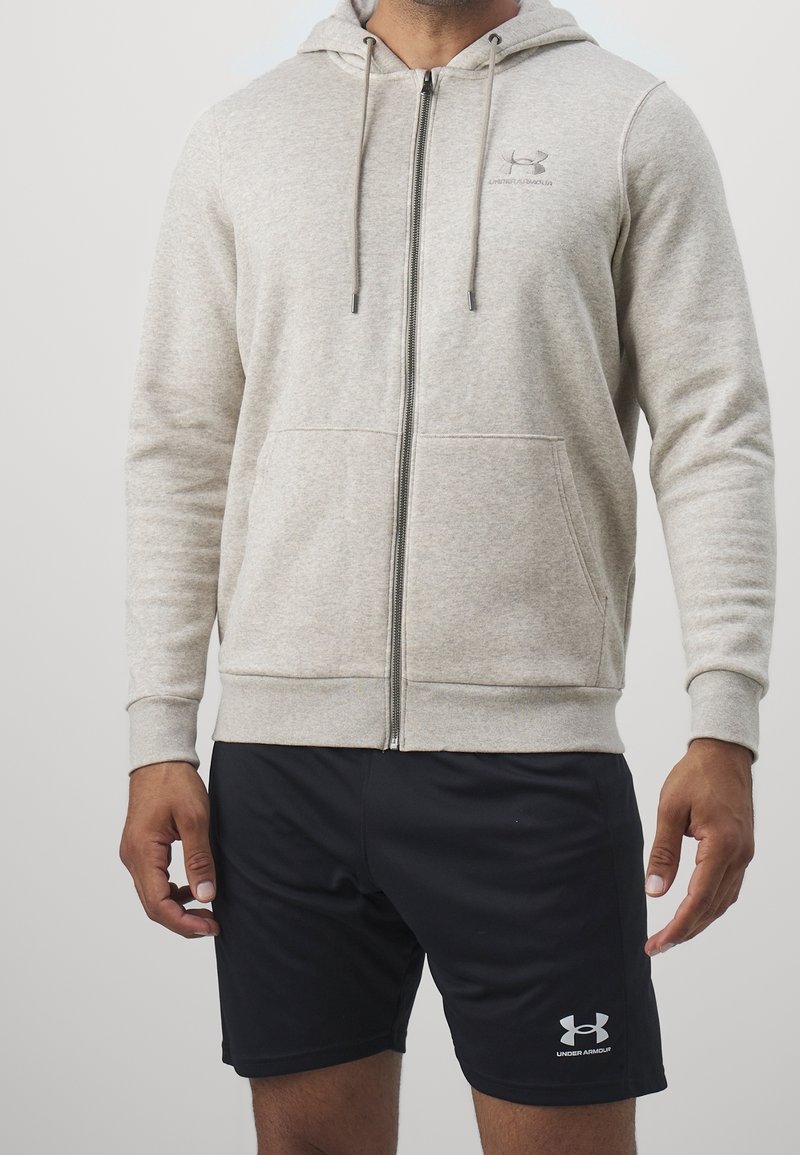 Homme portant un sweat à capuche gris clair zippé et un short noir Under Armour, debout devant un fond uni.