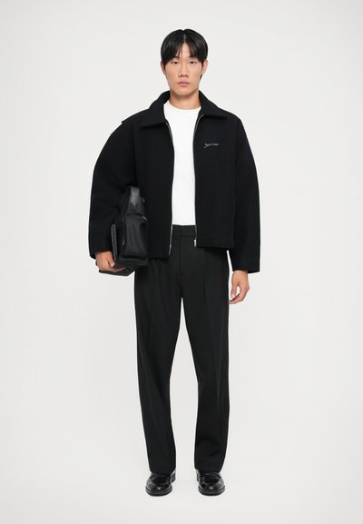 Schwarze Wolljacke mit Stehkragen, weißes T-Shirt, maßgeschneiderte schwarze Hose und schwarze Schuhe. Models halten eine schwarze Tasche.