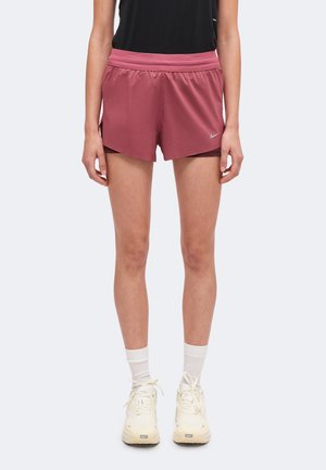 Osoba stojąca, ubrana w mauve sportowe szorty, czarną koszulkę, białe skarpetki i kremowe sneakersy, pokazana od pasa w dół.